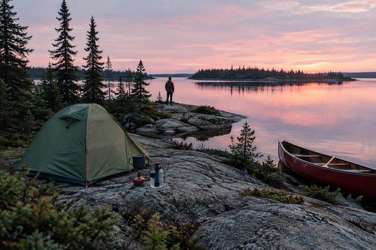 Small tent pitched on granite rock beside a calm boreal lake in Canada at dusk, with a canoe pulled up on shore and spruce trees behind