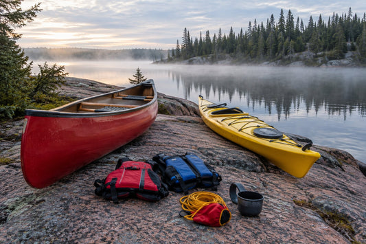 Red canoe pulled up on a granite shoreline of a calm boreal lake in Ontario, with paddles, PFDs and a coiled throw line resting on the rocks