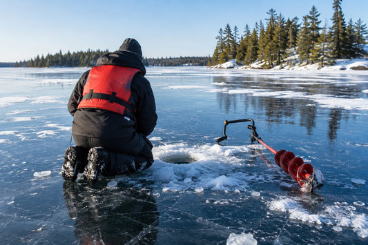 Person in a lifejacket kneeling on clear blue lake ice to measure a drilled test hole on a frozen boreal lake in Canada, with pine shoreline