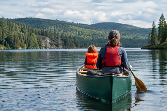 Child in a bright orange lifejacket with a head-support collar sits in a canoe on a calm Laurentian lake as an adult paddles at the stern